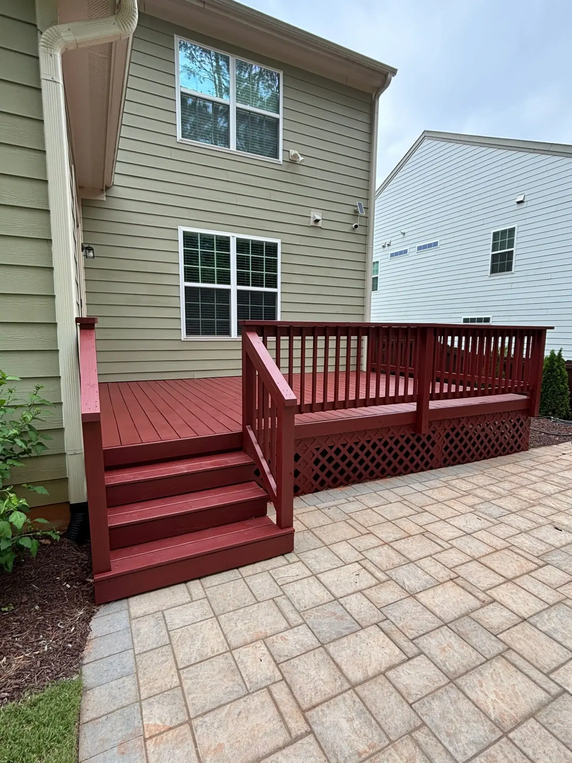 Vibrant Red Deck Painting and Staining Services A residential house with light green siding and a striking red painted wooden deck leading to a paved patio.