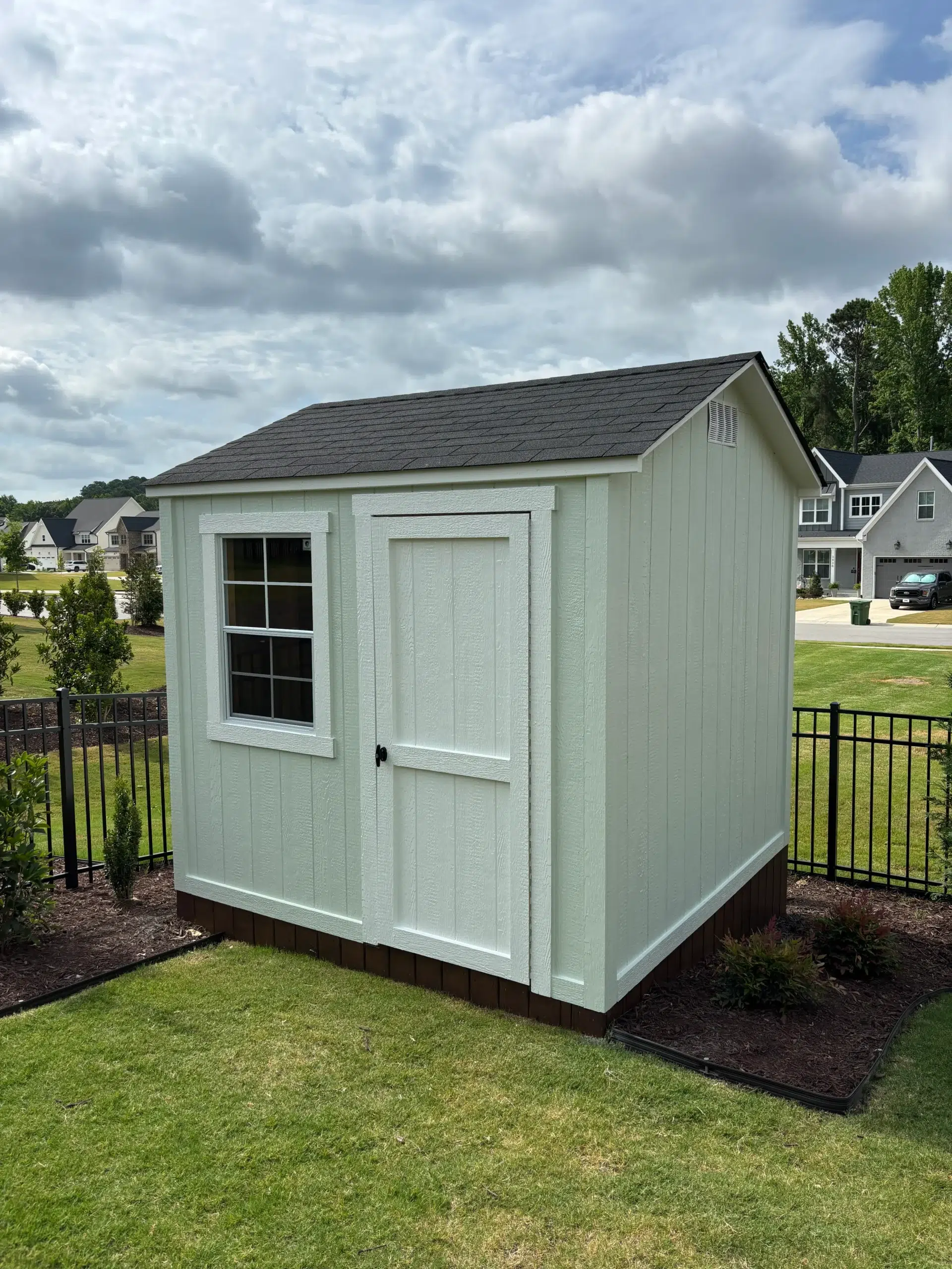Quality Shed Painting and Outdoor Structure Finishing A freshly painted light green backyard shed with white trim and a brown base in a sunny yard.
