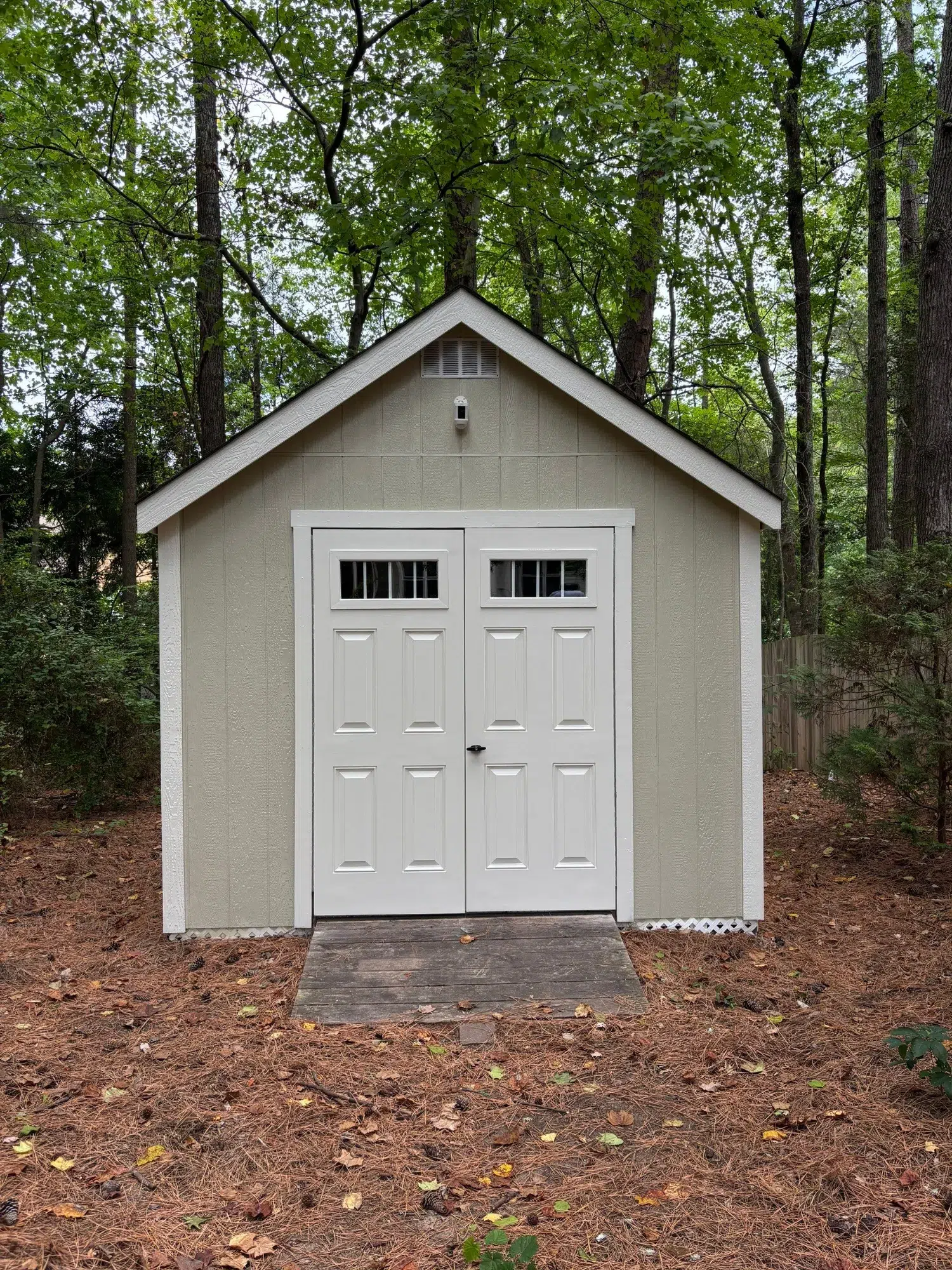 Painted Garden Shed Exterior Tan-painted fiber cement siding garden shed with white-trimmed double doors and peaked roof in wooded setting