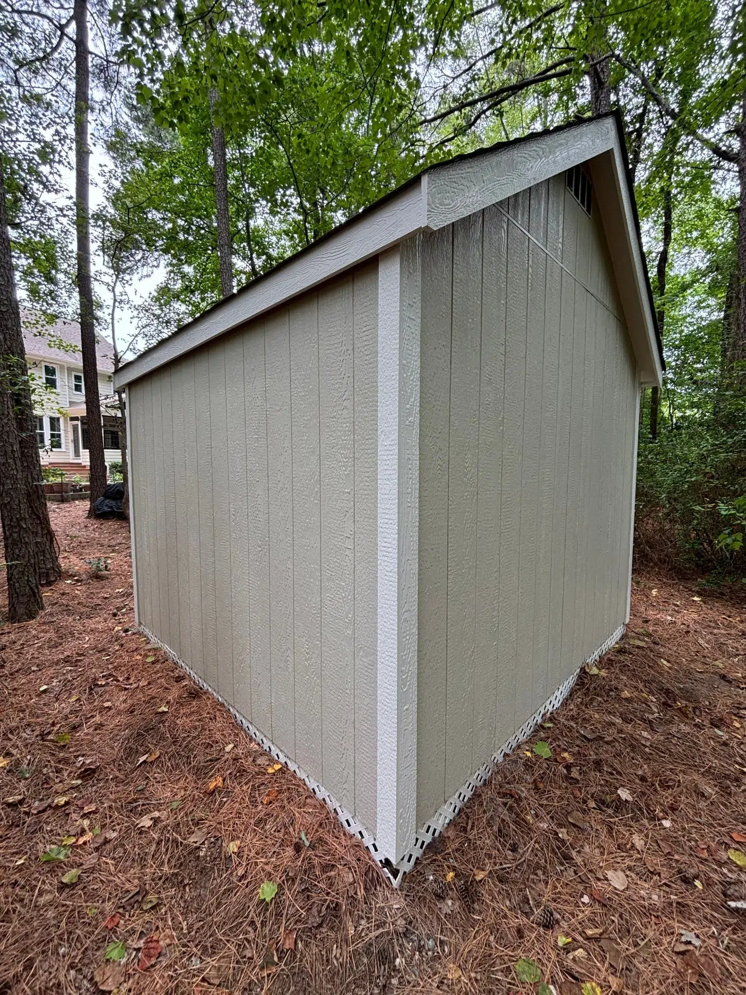 Tan Fiber Cement Garden Shed Tan-painted fiber cement siding garden shed with vertical wood-grain texture and dark roof in wooded backyard setting